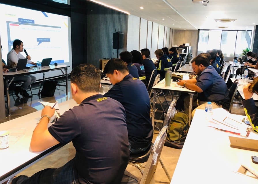 Trainees in SECCO polo shirts take notes in a classroom while an instructor presents slides