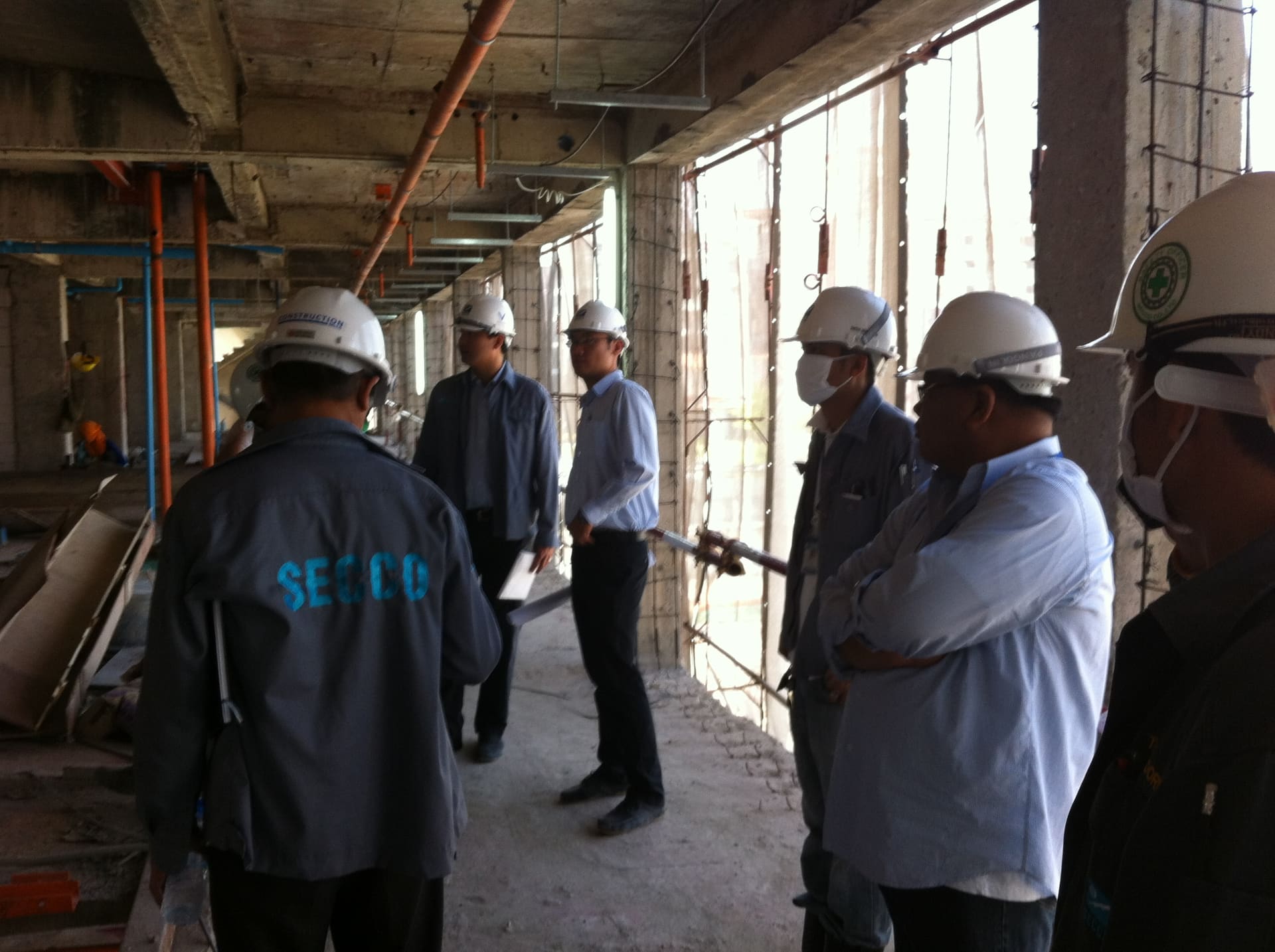 A group in white hard hats, one in a SECCO jacket, on an unfinished concrete floor under exposed ceiling pipework
