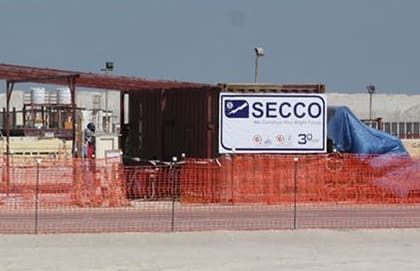 SECCO signboard on a site compound fence, in front of a shipping container and a steel canopy, behind orange mesh fencing