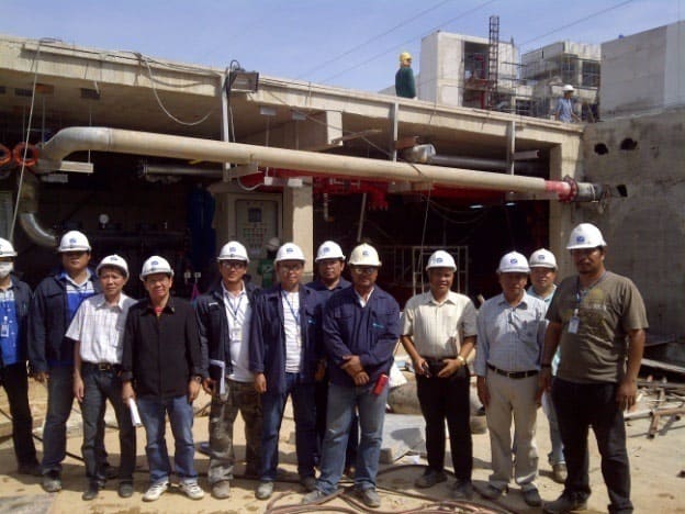 Group of men in white hard hats stand in front of a partly built concrete plant structure with large pipes