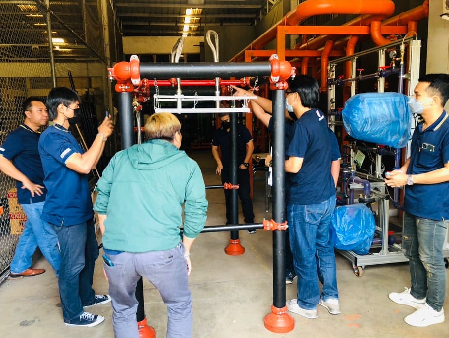 Trainees gather round a pipe-support training rig with orange-red grooved couplings, beside a valve test bench on wheels