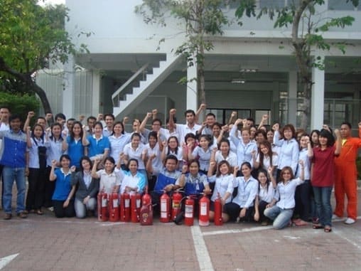 Large group of staff outside an office building with red fire extinguishers lined up in front of them