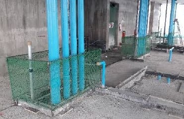 Green mesh barricade around the base of four blue riser pipes on a bare concrete floor