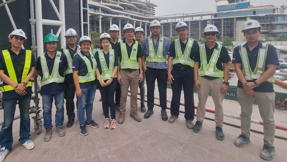 Twelve SECCO staff in hard hats and hi-vis vests on a paved deck, with scaffolding and multistorey buildings behind them
