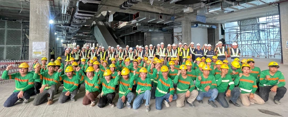 SECCO site team in green shirts and yellow hard hats kneeling with fists raised, with staff in white hard hats and hi-vis vests standing behind, inside a building under construction