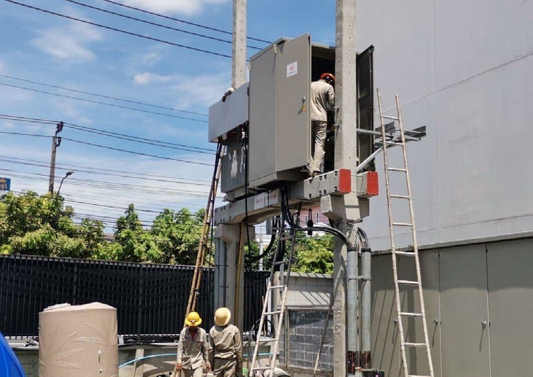 Workers in hard hats fit out a grey electrical enclosure on a two-pole concrete platform beside a building, with ladders against it