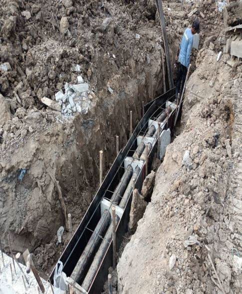 Deep excavated trench with conduits laid on white spacers between dark formwork panels, and a worker at the far end