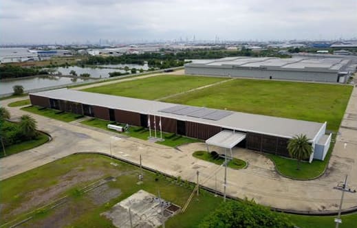Aerial view across lawns of a low building with a small rooftop solar array, a pond and an industrial area beyond