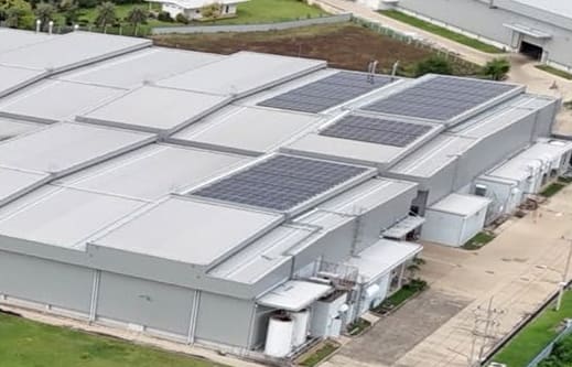 Aerial view of a large industrial building with solar panel arrays on its roofs and low white service annexes along one side