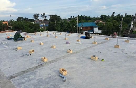 Workers set out mounting posts in small timber formwork boxes across a flat concrete roof, with trees and houses beyond