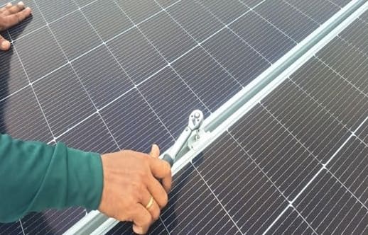 A worker's hand tightens a panel clamp on an aluminium rail with a ratchet, between two solar panels