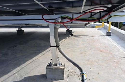 Underside of a solar array: a mounting post on a concrete footing, with grey flexible conduit and red, black and green-yellow cables