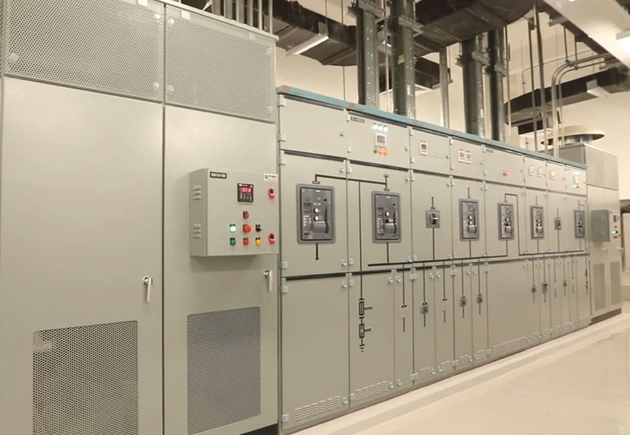 Line of grey switchgear panels with circuit-breaker doors under ductwork, pipes and conduit in an electrical room