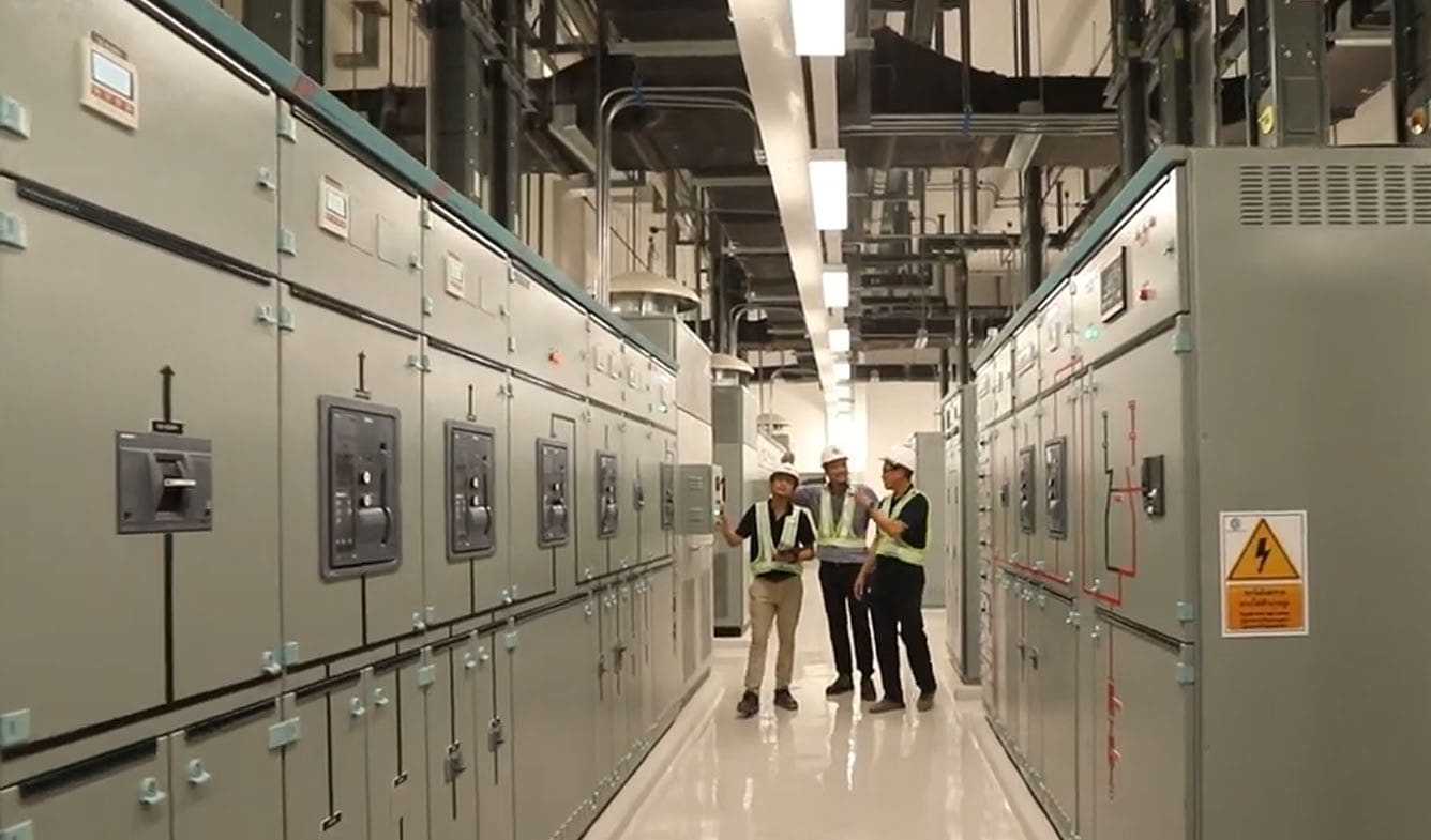 Three engineers in hard hats and hi-vis vests talk in an aisle between rows of grey switchgear panels