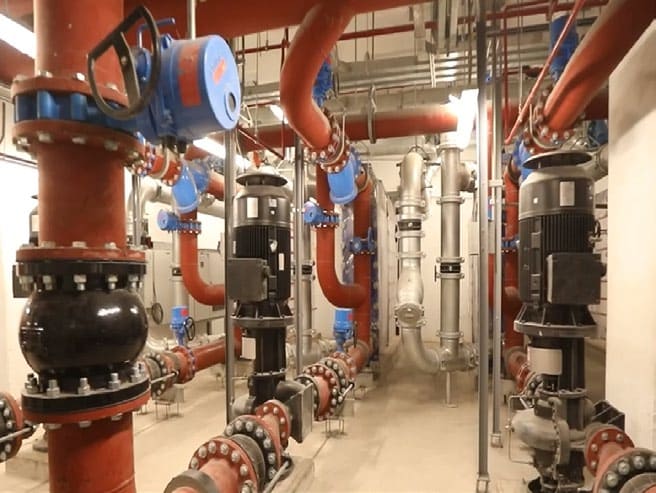 Pump room with vertical in-line pumps, red-painted pipework, blue motorised valve actuators and a black flexible connector