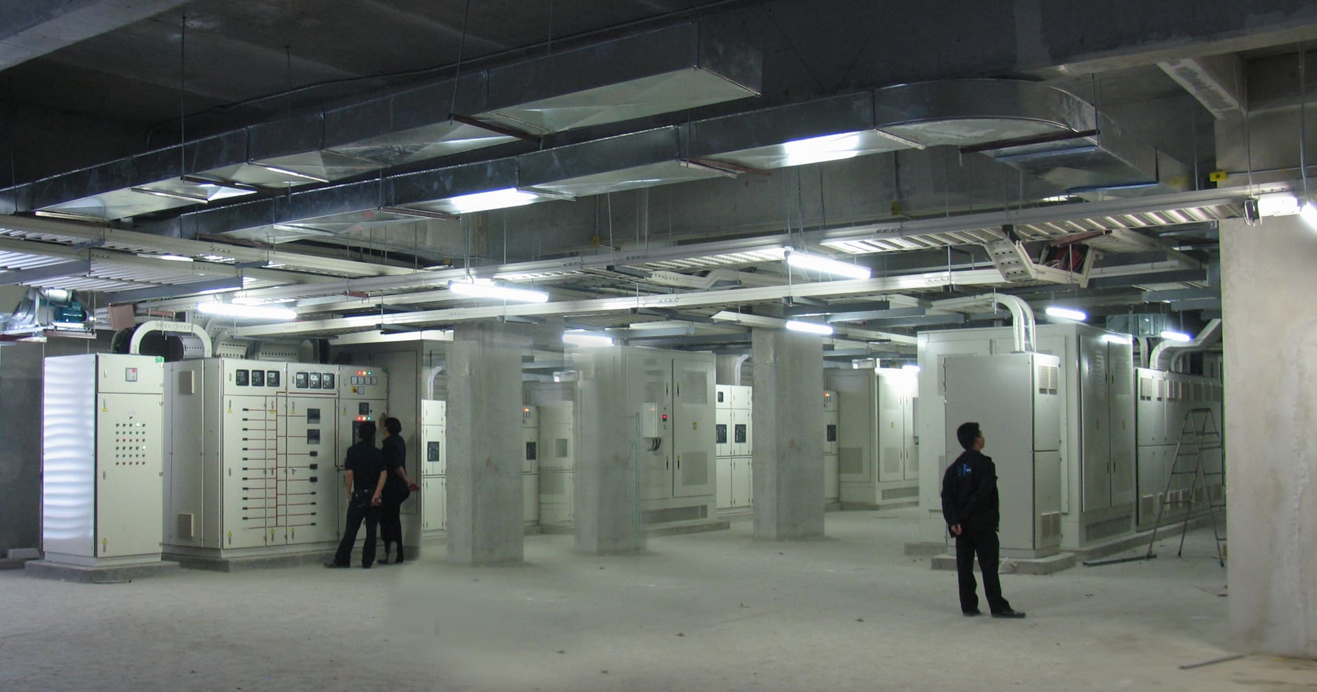Electrical room with rows of switchboard cabinets under ducts and cable trays, where two men inspect a panel