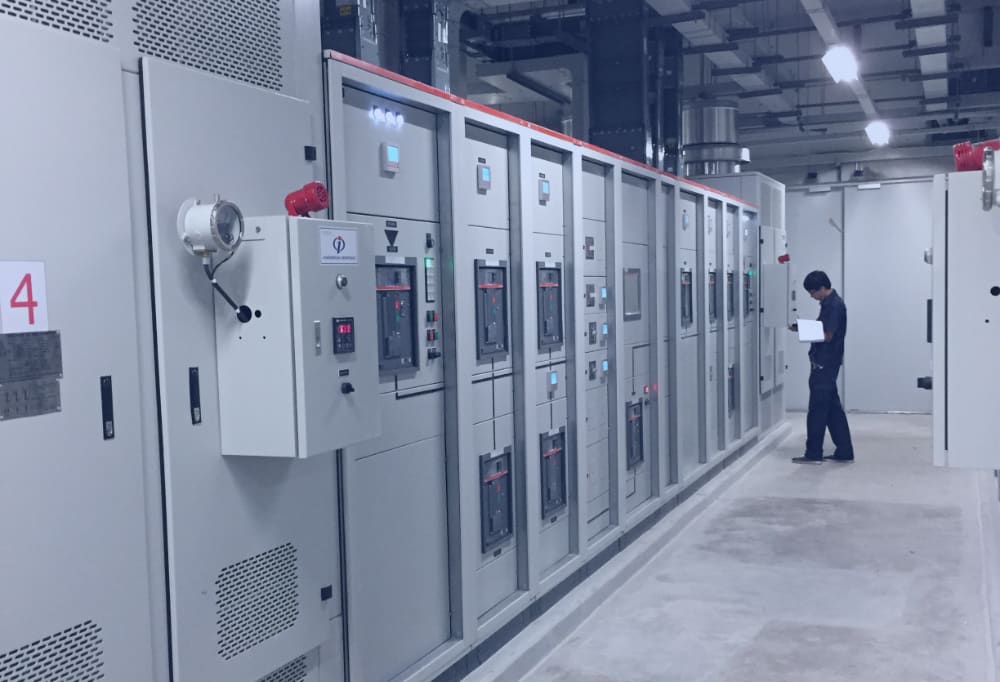A technician in dark workwear reads a folder beside a long row of grey switchgear cabinets in an electrical room