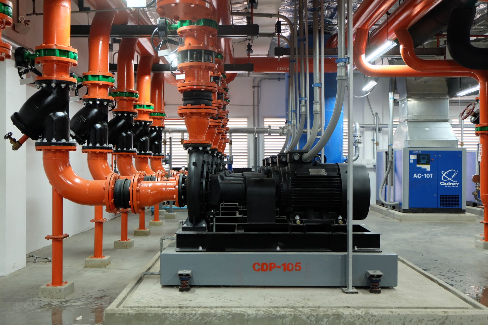 Row of condenser-water pumps with orange pipework and a blue air compressor in a plant room
