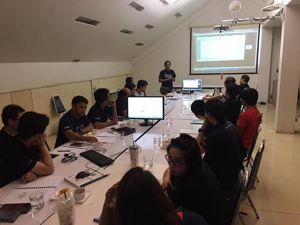 SECCO staff seated along a long table with computer monitors, keyboards and course booklets listen to a presenter in front of a projection screen