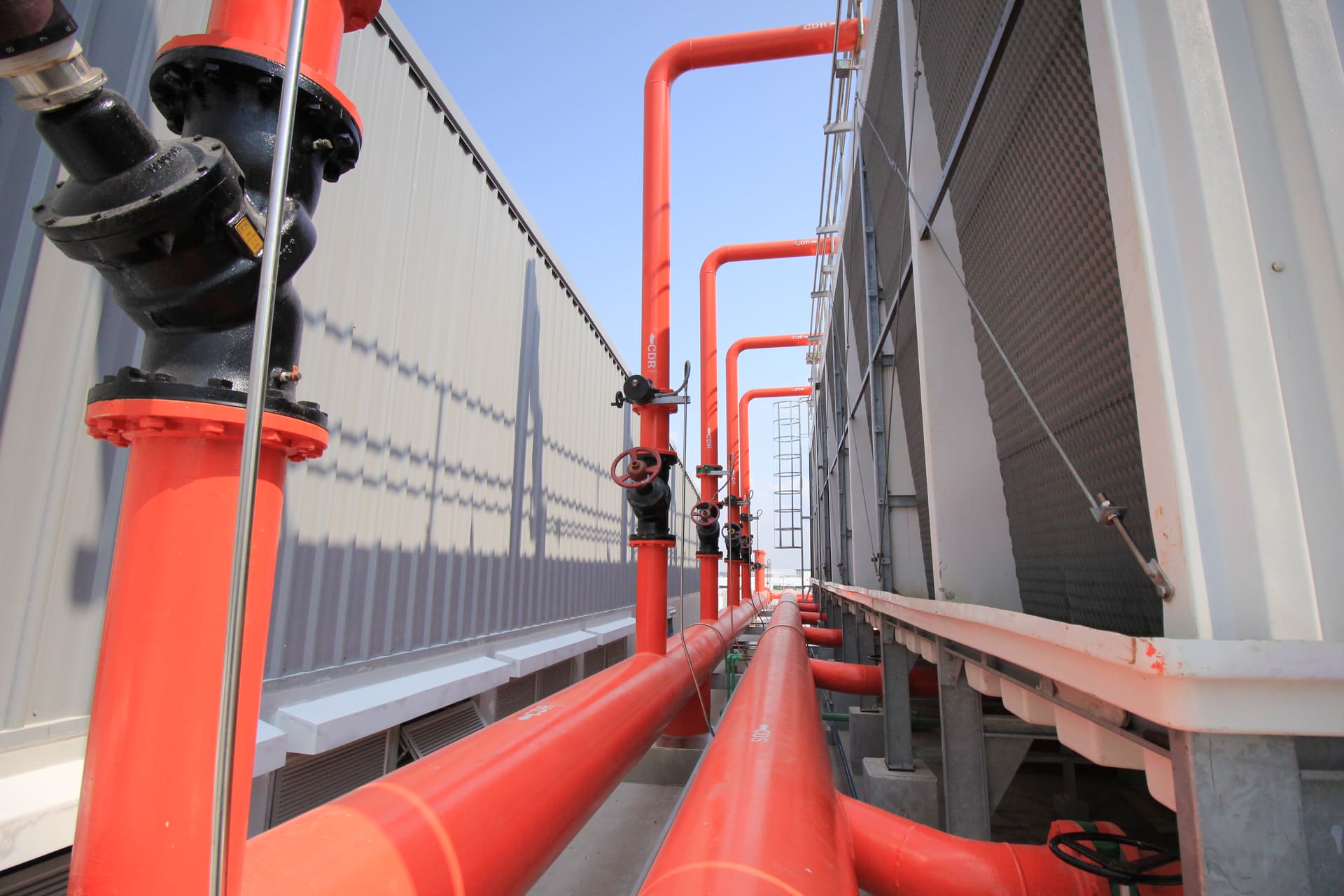 Orange-red condenser-water pipes with valves running between rows of rooftop cooling towers under a blue sky