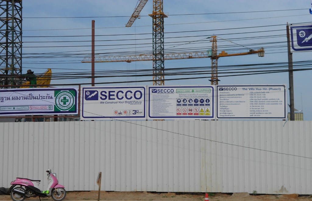 Construction site hoarding with SECCO banners and safety-rule boards, tower cranes behind