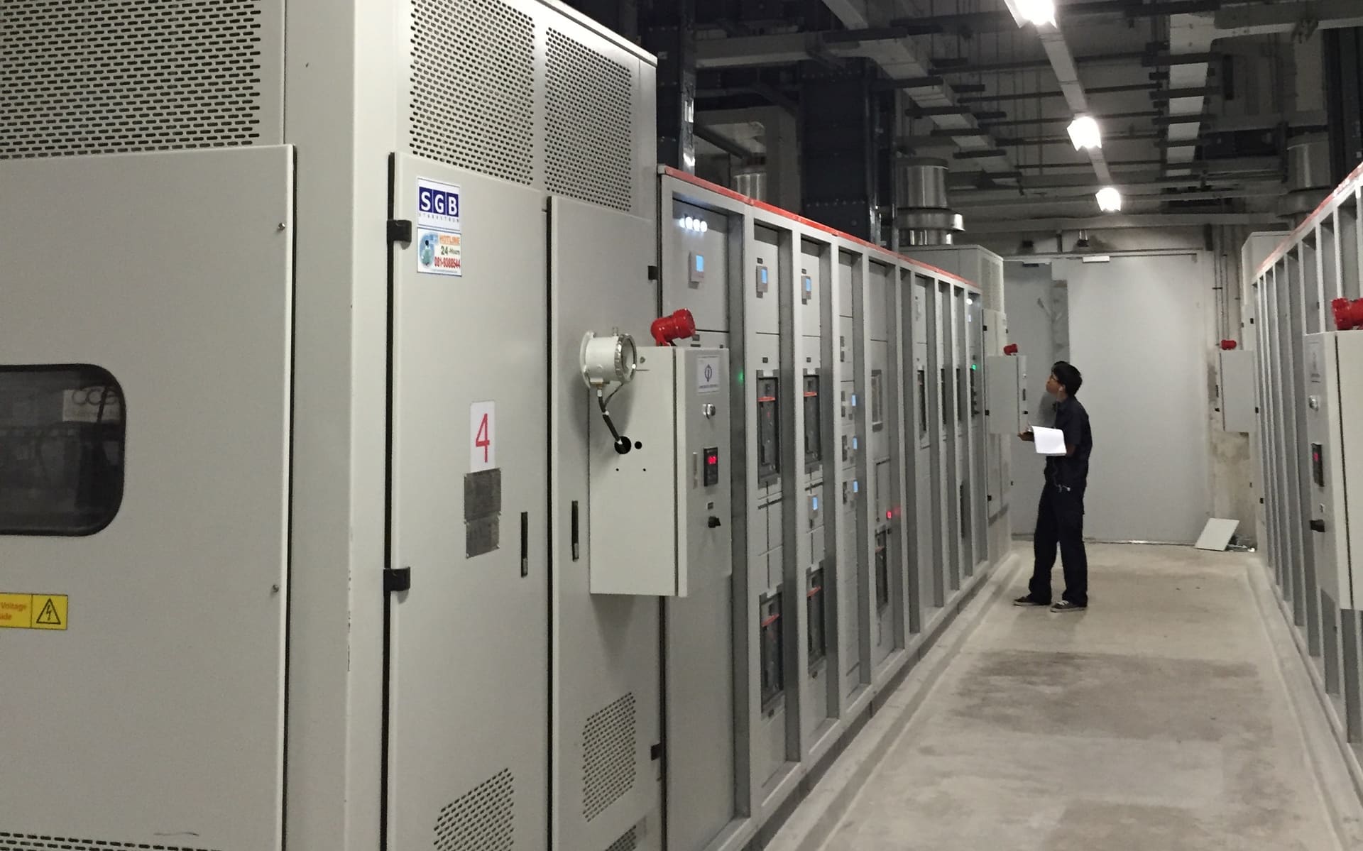 A worker holding papers looks up at a long row of grey switchgear cabinets in an electrical room