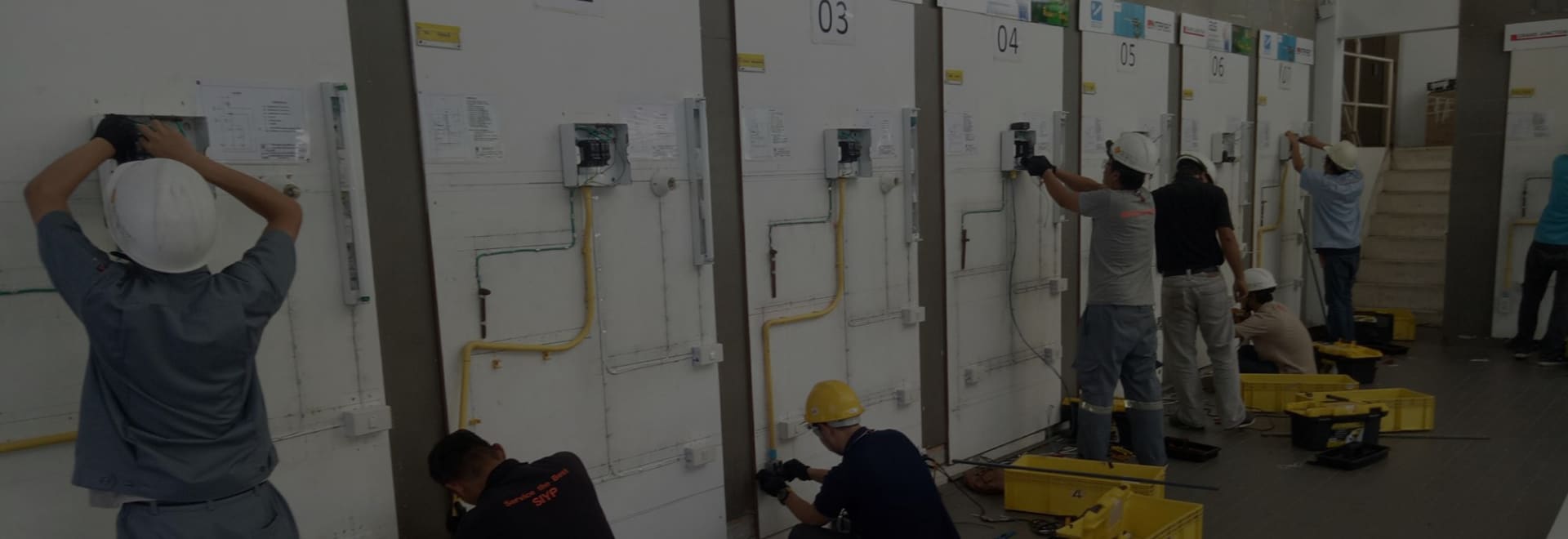 Candidates, most in hard hats, wiring circuits on a row of numbered white test boards, with tool crates on the floor
