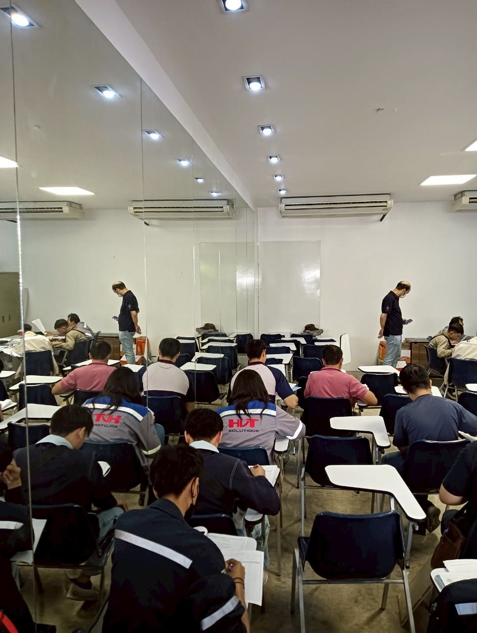 Candidates write on papers at tablet-arm chairs in a white classroom while a masked man stands at the back of the room