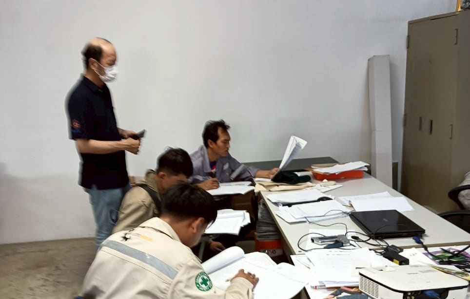 Candidates writing in booklets at a table with a projector and laptop while a masked man stands behind them