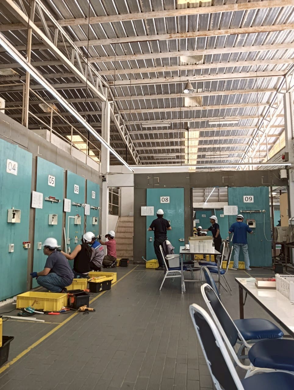 Wide view of a high-roofed workshop where candidates in hard hats wire rows of numbered teal practice panels