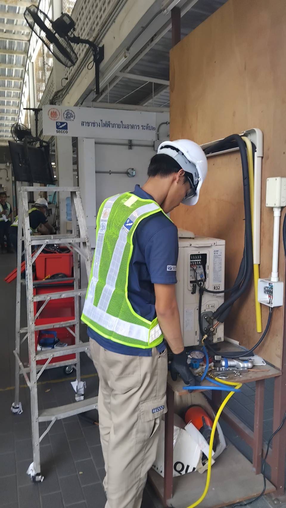 Test candidate in hard hat and hi-vis vest connects gauge hoses to an outdoor air-conditioning unit mounted beside a plywood test board