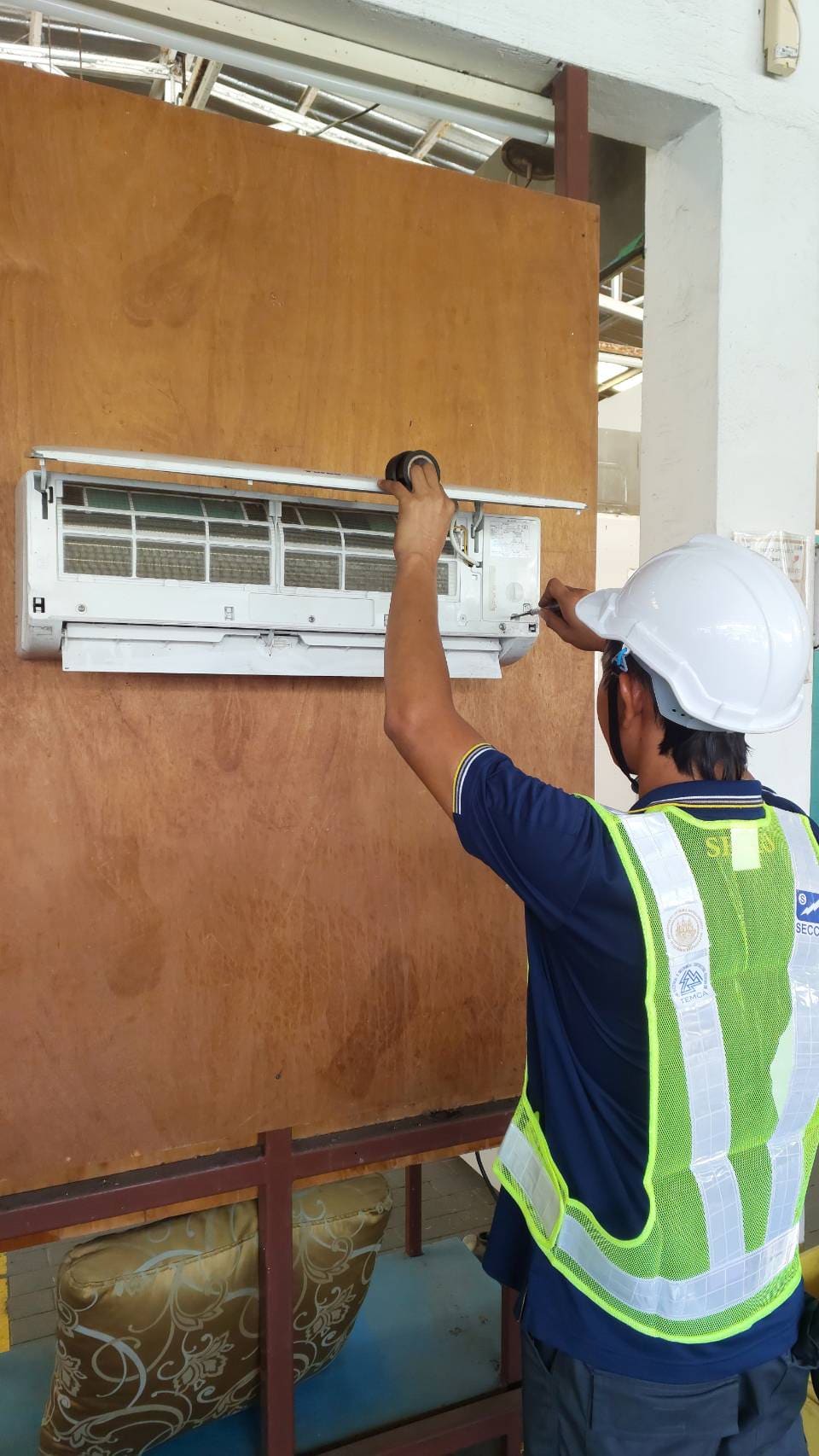 Test candidate in hard hat and hi-vis vest uses a screwdriver on an indoor air-conditioner unit whose front panel is raised