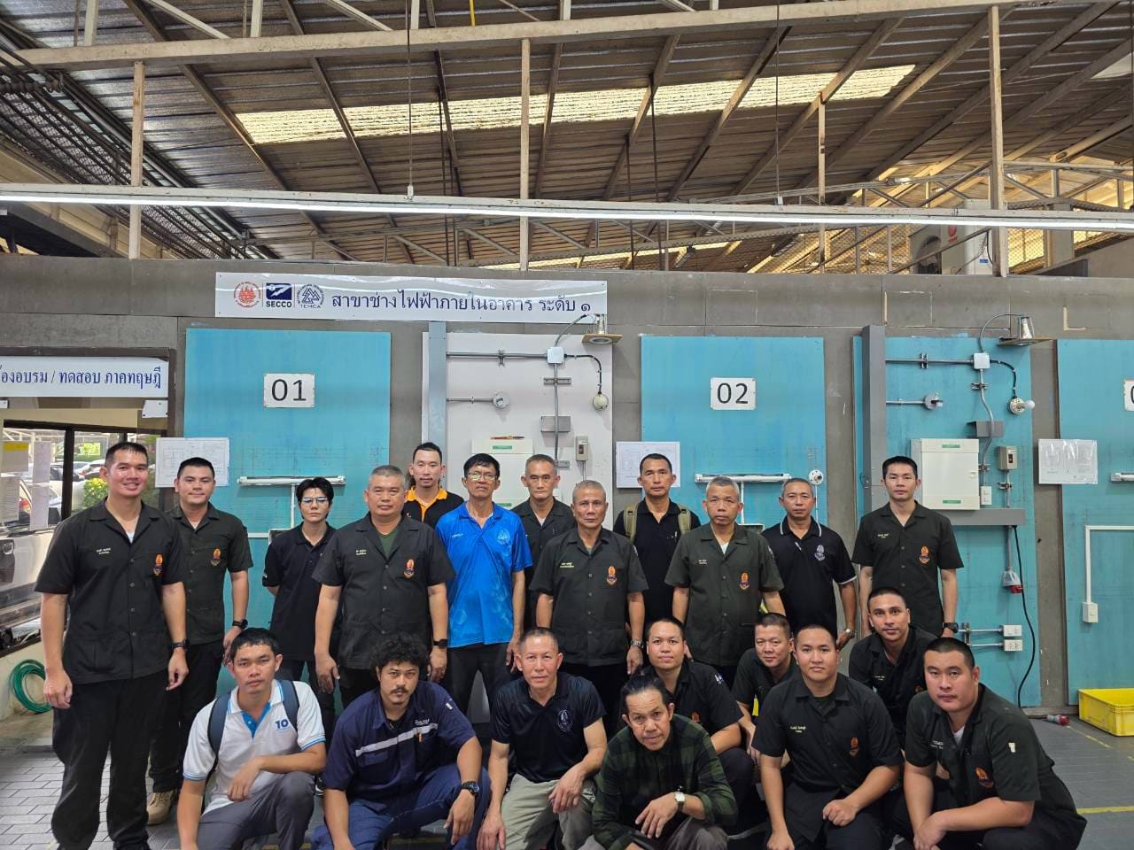 About twenty men pose in front of numbered blue electrical test booths under a banner for building electrician level 1