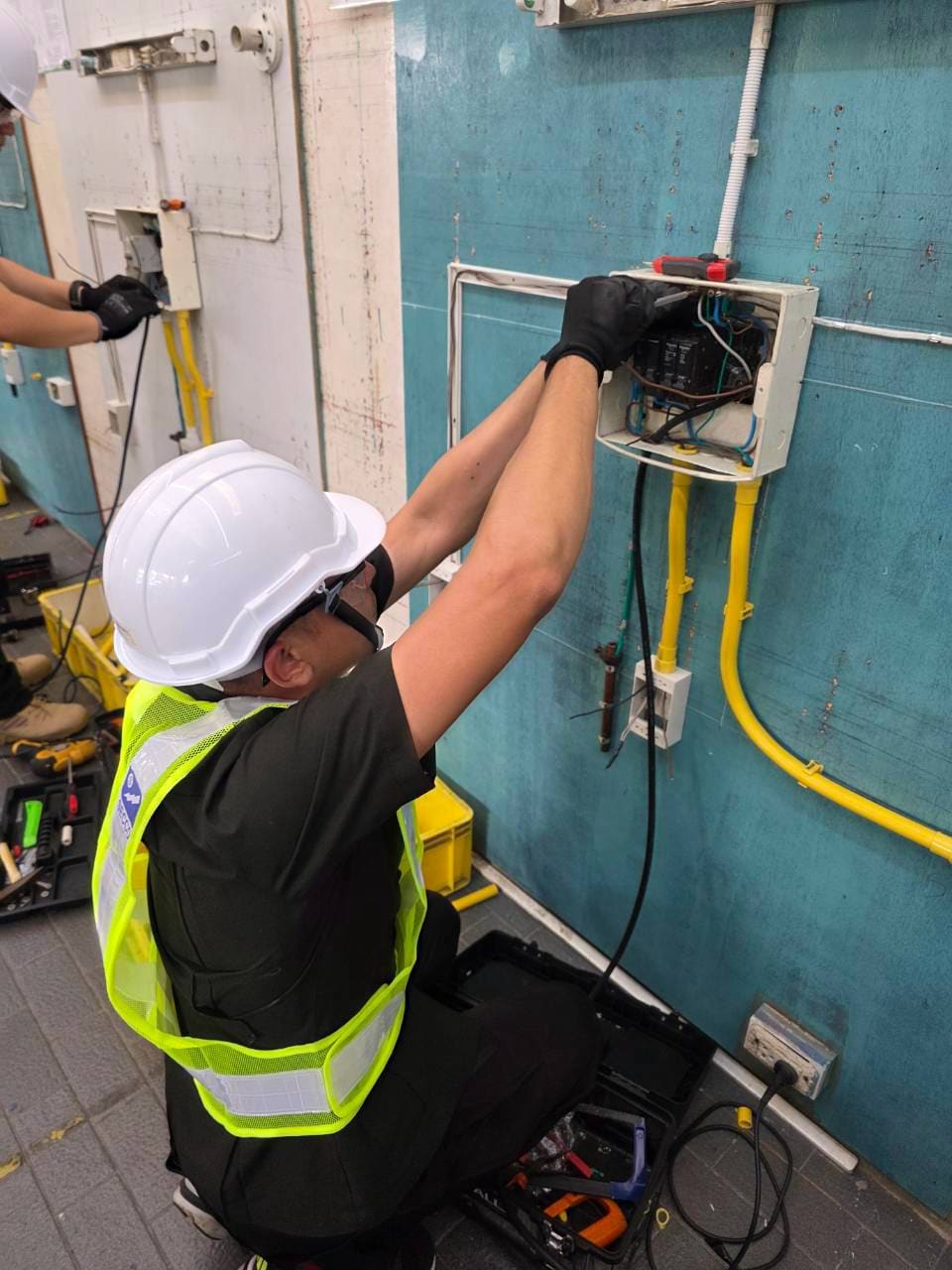 Test candidate in hard hat and hi-vis vest reaches up to wire a consumer unit on a blue test board