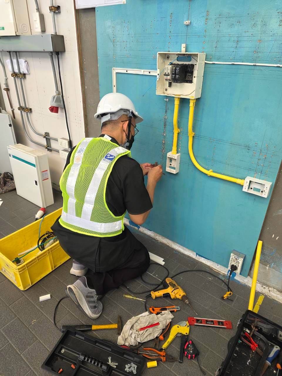 Test candidate in a face mask kneels to fix a small metal fitting beside an empty switch box; drill, pliers and toolboxes on the floor