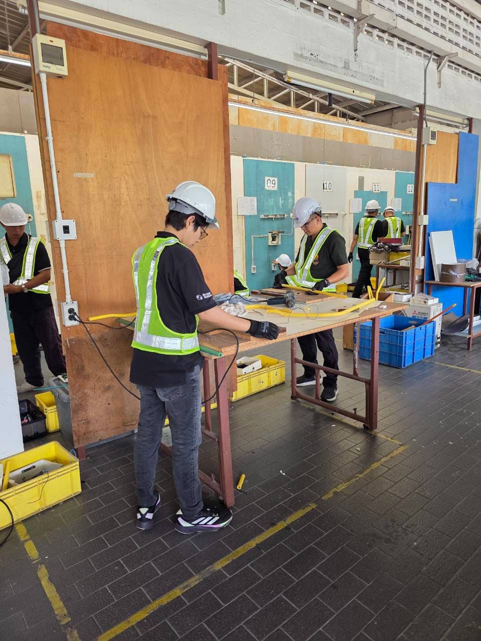 Test candidates in hard hats and hi-vis vests work with yellow conduit at a workbench in an open-sided workshop