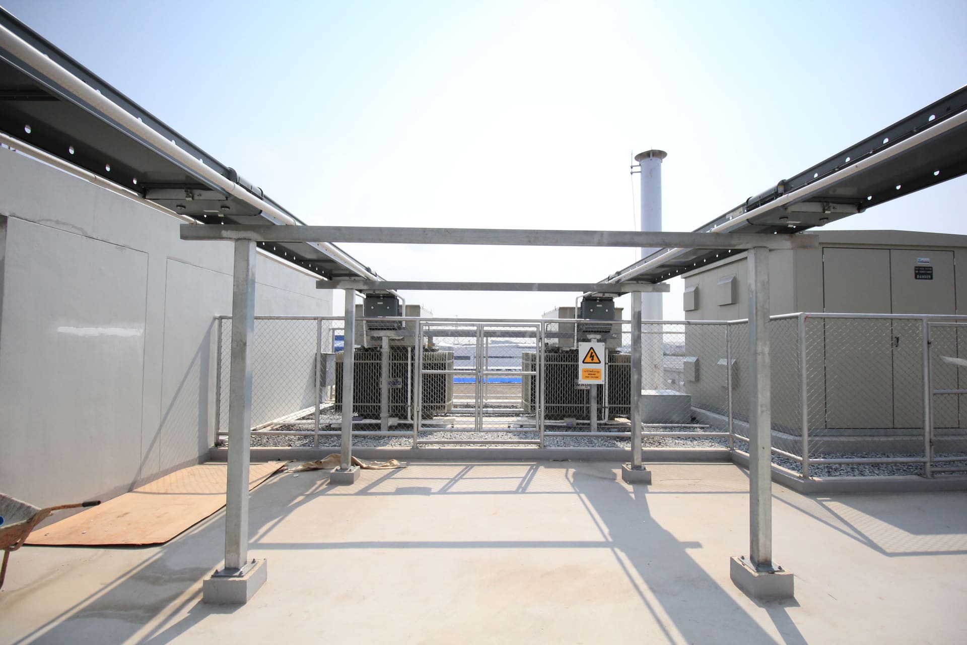 Rooftop yard with two fenced transformers under steel cable-tray gantries, with a high-voltage warning sign and a tall metal stack