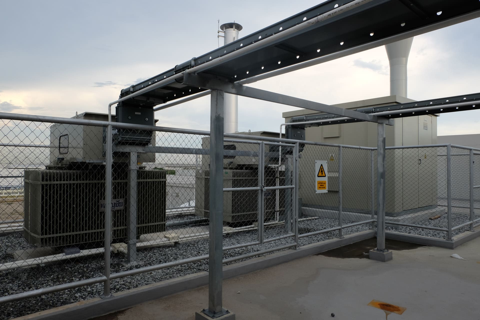 Fenced rooftop transformer yard under a steel cable-tray gantry, with a high-voltage warning sign